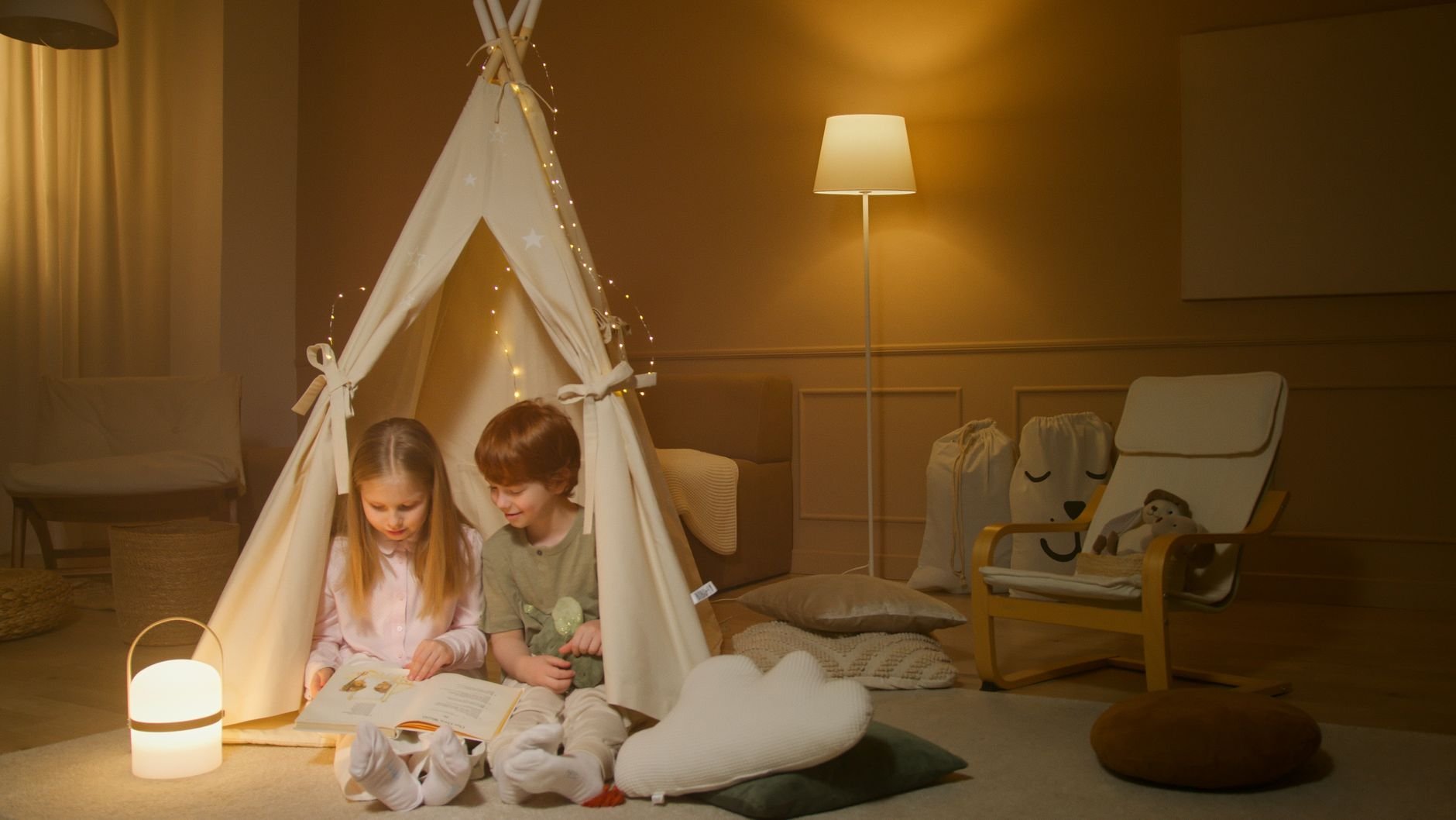 Two children reading a book in a warmly lit indoor tent, creating a cozy atmosphere.